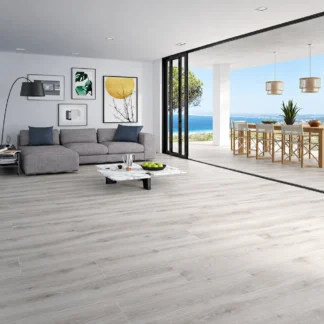 Algord Grey Porcelain Tile with a gray wood-look finish, installed in a bright modern living room featuring a gray sofa, floor-to-ceiling windows, and an ocean-view terrace.